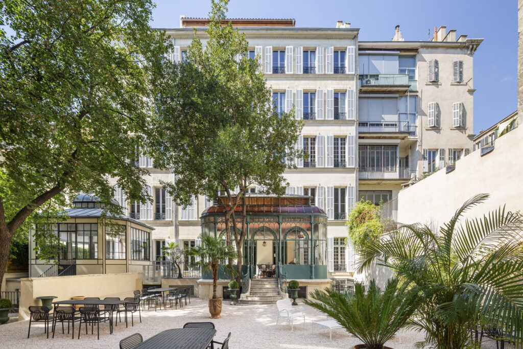 Sunlit courtyard with café-style black tables and chairs on pale gravel, framed by leafy trees and potted palms, in front of a tall cream-and-white apartment building with shuttered windows, plus two glass conservatories, one a green ornate pavilion reached by steps.